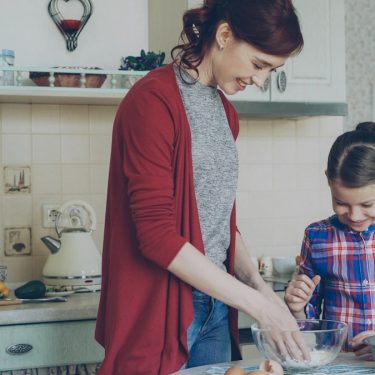 Little cheerful girl helping her mother in the kitchen mixing dough for cookies. Family, food, home and people concept