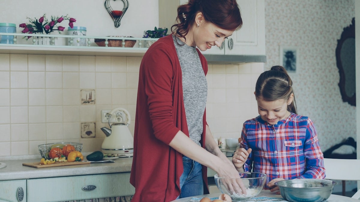 Little cheerful girl helping her mother in the kitchen mixing dough for cookies. Family, food, home and people concept